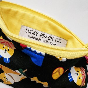 May include: A black zippered pouch with a yellow lining and a white label that reads "LUCKY PEACH CO handmade with love". The pouch is decorated with a repeating pattern of cartoon dogs wearing hats and scarves.