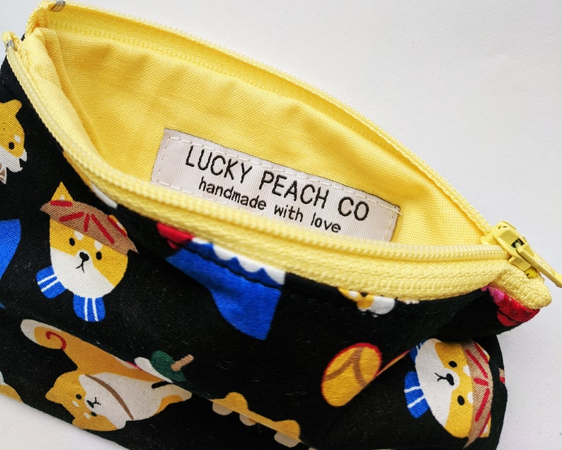 May include: A black zippered pouch with a yellow lining and a white label that reads "LUCKY PEACH CO handmade with love". The pouch is decorated with a repeating pattern of cartoon dogs wearing hats and scarves.