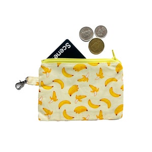 May include: A small yellow zippered pouch with a banana print. The pouch has a silver clip and a black card inside. Three silver coins are on top of the pouch.