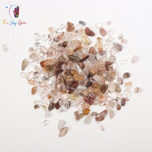 May include: A pile of small, clear, brown, and red quartz crystals. The crystals are scattered on a white surface.