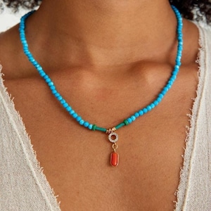 May include: A turquoise beaded necklace with a gold pendant featuring a small circle with clear stones and a rectangular red stone.