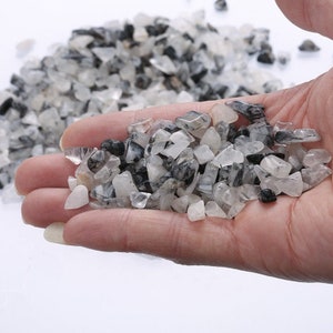 May include: A handful of small, irregularly shaped, clear and opaque crystals with black inclusions. The crystals are held in an open hand, with a white background.