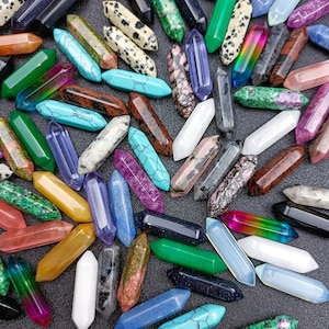 May include: A collection of colourful, pointed crystal wands in various shades of blue, green, purple, red, orange, yellow, brown, white, and black. The wands are made of natural stone and are approximately 5 cm long.
