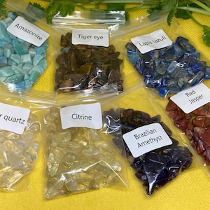 Könnte beinhalten: Sieben kleine Beutel mit geschliffenen Edelsteinen, jeweils mit der Art des Steins beschriftet: Amazonit, Tigerauge, Lapislazuli, Klarer Quarz, Citrin, Brasilianischer Amethyst und Roter Jaspis.