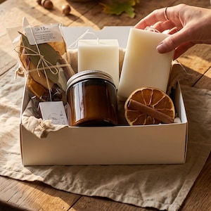 May include: A white gift box containing two rectangular off-white pillar candles, a small amber glass jar with a silver lid, a dried orange slice, a cinnamon stick, and a clear plastic bag filled with dried leaves and a small white label.