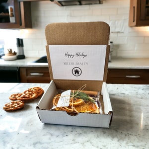 Può includere: Una piccola scatola di cartone con un coperchio bianco che dice "Buone Feste" e "Miller Realty" con un'icona di casa. La scatola è aperta e contiene un sacchetto di plastica trasparente con fette di arancia essiccate, bastoncini di cannella e rametti di sempreverde.