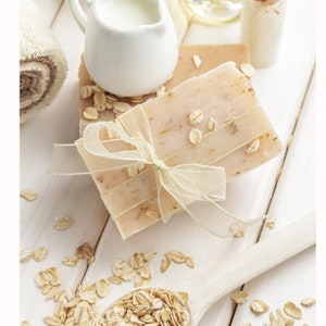 Puede incluir: Dos barras de jab&oacute;n de avena atadas con una cinta blanca sobre una superficie de madera blanca con copos de avena esparcidos.