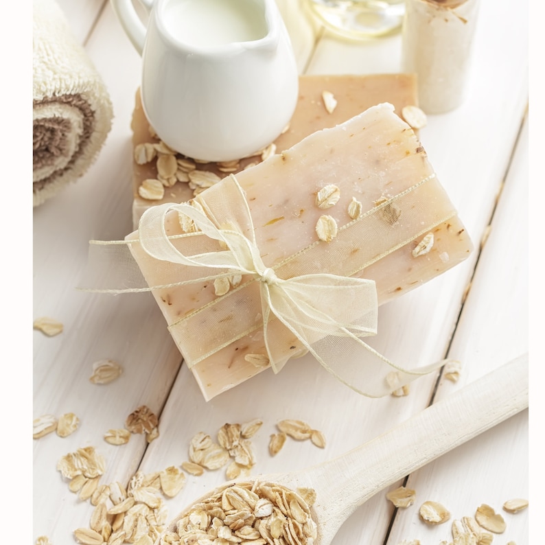 Puede incluir: Dos barras de jab&oacute;n de avena atadas con una cinta blanca sobre una superficie de madera blanca con copos de avena esparcidos.