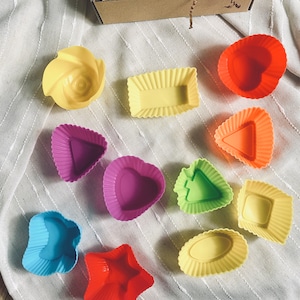 Puede incluir: Un conjunto de 11 moldes para hornear de silicona de colores en varias formas, incluyendo un coraz&oacute;n, una estrella, una flor y un rect&aacute;ngulo. Los moldes est&aacute;n sobre un fondo de tela blanca.