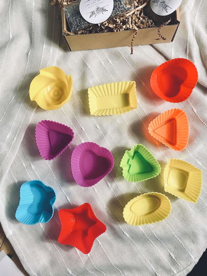Puede incluir: Un conjunto de 11 moldes para hornear de silicona de colores en varias formas, incluyendo un coraz&oacute;n, una estrella, una flor y un rect&aacute;ngulo. Los moldes est&aacute;n sobre un fondo de tela blanca.