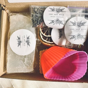 Puede incluir: Una caja de cart&oacute;n marr&oacute;n sobre un fondo blanco. La caja contiene una barra de jab&oacute;n blanca, lavanda seca, rodajas de naranja, una peque&ntilde;a botella de aceite y dos moldes de silicona. La caja tambi&eacute;n tiene tres pegatinas redondas con el texto "She and Brie" y las palabras "Dried Lavender", "Orange and Lavender Oil" y "Olive Soap Bar".