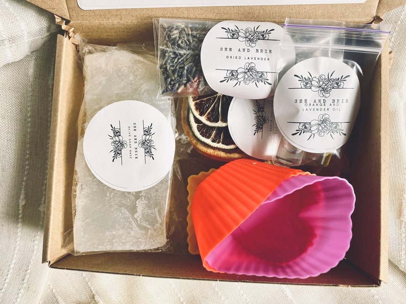 Puede incluir: Una caja de cart&oacute;n marr&oacute;n sobre un fondo blanco. La caja contiene una barra de jab&oacute;n blanca, lavanda seca, rodajas de naranja, una peque&ntilde;a botella de aceite y dos moldes de silicona. La caja tambi&eacute;n tiene tres pegatinas redondas con el texto "She and Brie" y las palabras "Dried Lavender", "Orange and Lavender Oil" y "Olive Soap Bar".