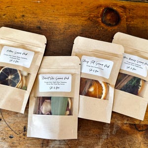 May include: Four kraft paper pouches with clear windows, each containing dried botanicals. The pouches have white labels with text. Visible contents include dried orange slices, cinnamon sticks, and other spices. The pouches are arranged on a wooden surface.