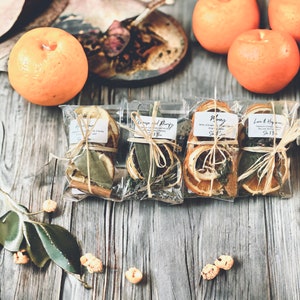 May include: Four clear cellophane packages tied with raffia, each containing dried herbs and spices. The packages are labeled with handwritten text. Oranges and other decorative items are arranged on a weathered wooden surface.