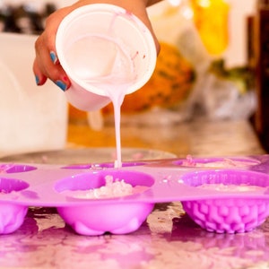 Puede incluir: Una persona vierte l&iacute;quido rosa en un molde de silicona morado con m&uacute;ltiples cavidades. El molde est&aacute; sobre una superficie plateada.