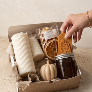 May include: A gift box containing two white ribbed pillar candles, a small beige pumpkin-shaped candle, a glass jar with a metal lid, a bag of dried orange slices labeled "Orange & Cinnamon", and a yellow wax tag with a monstera leaf design.
