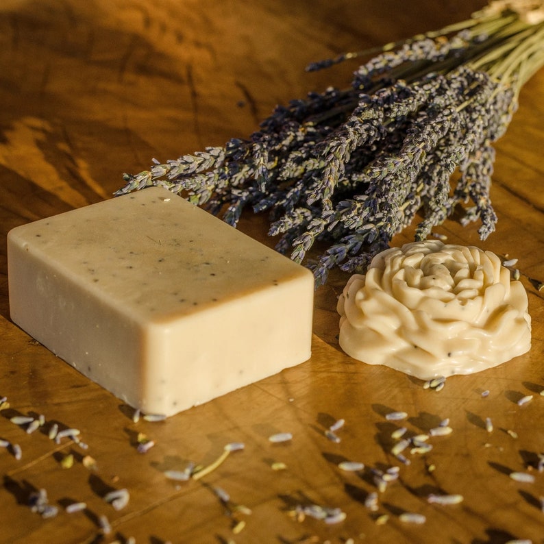 Puede incluir: Dos barras de jab&oacute;n hechas a mano, una con forma de flor y otra rectangular, ambas de color beige claro y textura moteada. Un ramo de flores de lavanda secas est&aacute; en el fondo.