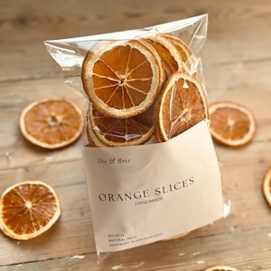 Puede incluir: Una bolsa de plástico transparente llena de rodajas de naranja secas. La bolsa tiene una etiqueta blanca con el texto "She & Brie ORANGE SLICES HANDMADE SET OF 21 NATURAL FRUIT HANDMADE IN SAN FRANCISCO".