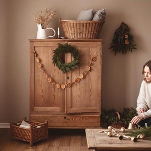 Puede incluir: Un interior acogedor con un armario de madera adornado con una corona verde y una guirnalda de rodajas de naranja secas. Una mujer está sentada cerca, rodeada de decoraciones navideñas, incluyendo campanas y vegetación. También se ven una cesta de mimbre y una jarra blanca.