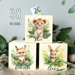 Könnte beinhalten: Drei Holzspardosen mit Tierillustrationen. Die Boxen zeigen ein Löwenjunges, einen Fuchs und ein Kaninchen, jedes umgeben von grünem Laub. Der Text "38 DESIGNS" steht über den Boxen. Die Löwenbox hat einen 20-Euro-Schein im Schlitz.