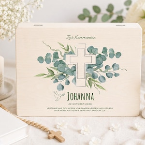 Puede incluir: Una caja de madera clara con un diseño de cruz y floral, el texto "Zur Kommunion" y el nombre "Johanna". La caja está decorada con hojas de eucalipto y una paloma. También hay una vela, una biblia y una pequeña figura de ángel.