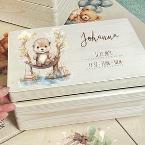 Könnte beinhalten: Helle Holzkiste mit einer Aquarellillustration eines Otters in einer Hängematte. Die Kiste ist mit dem Namen "Johanna" und Geburtsdaten personalisiert: 16.12.2023, 12:32, 3550g, 56cm.