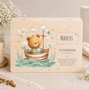 May include: A rectangular wooden box featuring a printed illustration of a brown bear in a small boat on water, accompanied by two white doves, clouds, and stars. The text reads Marcus, Zur Kommunion, 07 October 2025, and a German Bible verse.