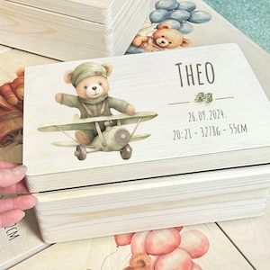 Puede incluir: Una caja de madera para recuerdos con un acabado de madera clara. La tapa presenta una ilustración de dibujos animados de un oso de peluche con un gorro de piloto y volando un avión. La caja está personalizada con el nombre "Theo", la fecha "26.09.2024" y la hora y las medidas "20:21 - 3278G - 55CM".
