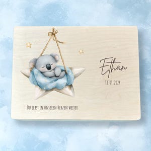 Peut inclure: Une plaque rectangulaire en bois avec une illustration aquarelle d'un koala endormi enveloppé dans une couverture bleue, reposant sur une étoile blanche suspendue par une corde. Le texte indique Ethan, 13.01.2024, et Du lebst in unseren Herzen weiter.