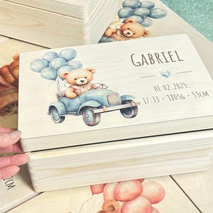 Può includere: Scatola ricordo in legno con un'illustrazione di un'auto azzurra. Un orsacchiotto guida l'auto e tiene in mano dei palloncini blu. La scatola è personalizzata con il nome "Gabriel", data di nascita, ora, peso e altezza (53 cm).