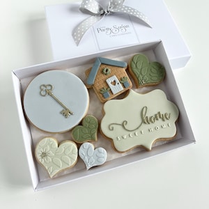 Op de afbeelding: Een witte cadeaudoos met een grijs gestippeld lint dat in een strik is gebonden. De doos bevat een set van zes gedecoreerde koekjes. De koekjes zijn versierd met een huis, een sleutel, harten en de woorden "Sweet Home".