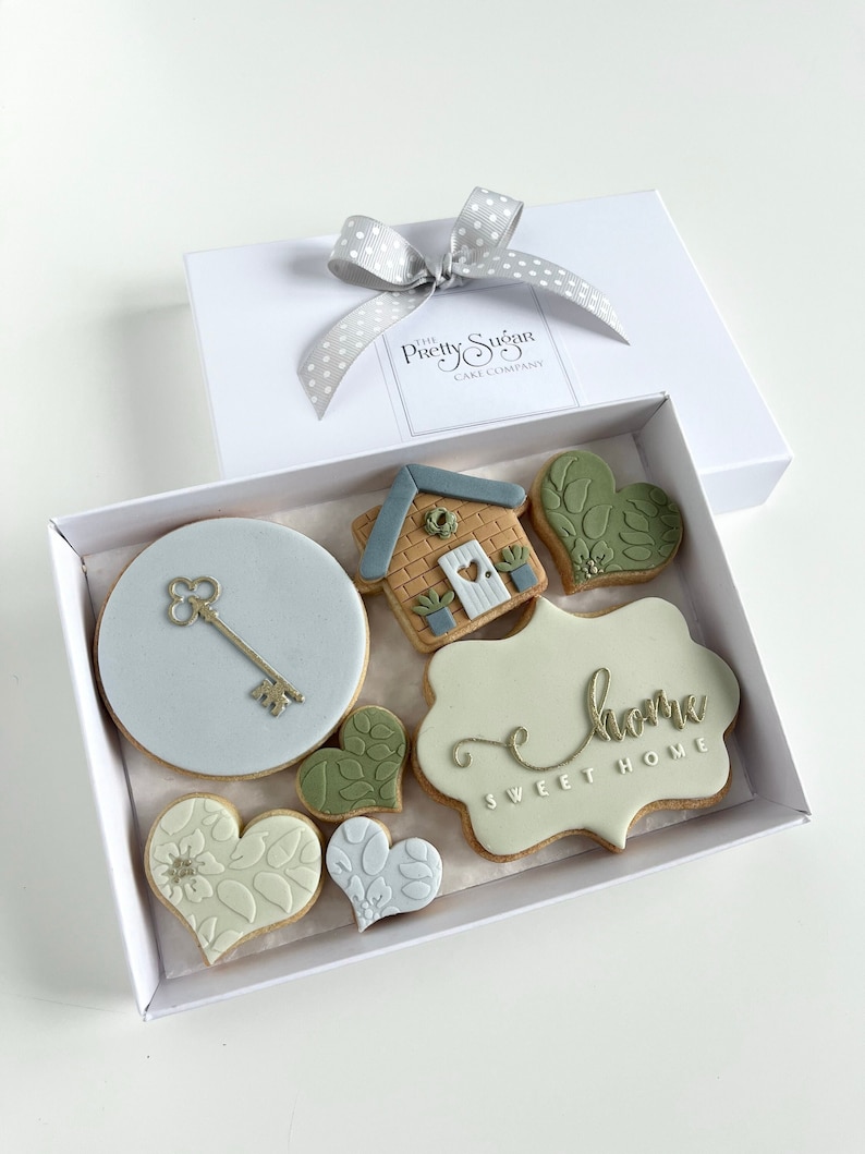 Op de afbeelding: Een witte cadeaudoos met een grijs gestippeld lint dat in een strik is gebonden. De doos bevat een set van zes gedecoreerde koekjes. De koekjes zijn versierd met een huis, een sleutel, harten en de woorden "Sweet Home".