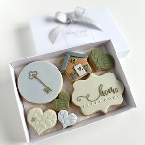 Op de afbeelding: Een witte cadeaudoos met een zilveren lint en een set van 8 gedecoreerde koekjes erin. De koekjes zijn versierd met een huis, een sleutel, harten en de woorden "home sweet home".