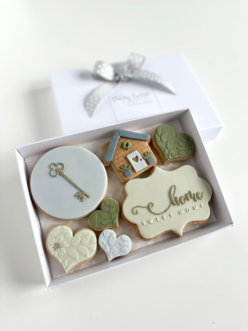 Op de afbeelding: Een witte cadeaudoos met een zilveren lint en een set van 8 gedecoreerde koekjes erin. De koekjes zijn versierd met een huis, een sleutel, harten en de woorden "home sweet home".