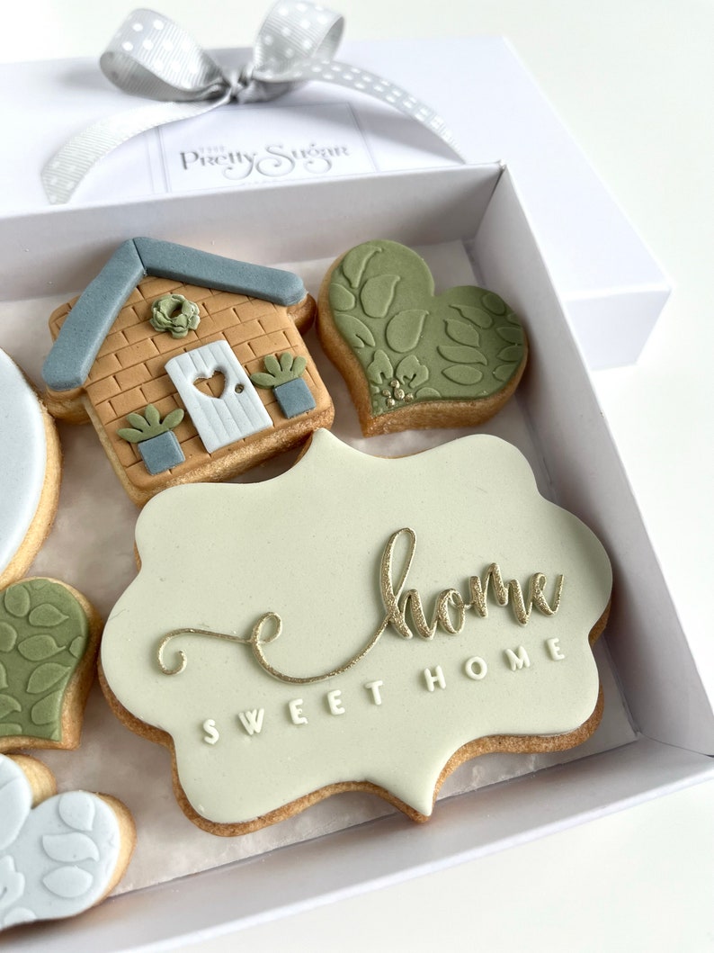Op de afbeelding: Een set gedecoreerde koekjes in een witte doos. De koekjes zijn versierd met een huis, harten en de woorden "Sweet Home".