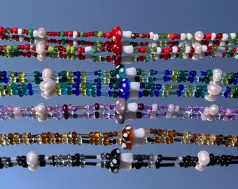 May include: Five beaded necklaces with mushroom pendants and various coloured beads. The necklaces feature red, green, white, blue, purple, brown, and black beads. Some necklaces have pearl accents. The background is a reflective surface with a blue sky.