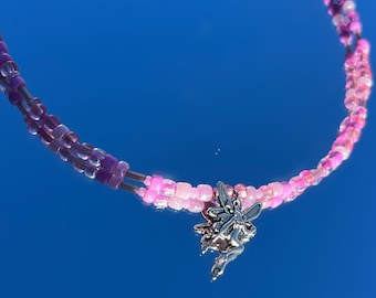 May include: A beaded necklace with pink and purple beads. The necklace features a silver fairy charm. The beads are arranged in two strands. The background is a solid blue.