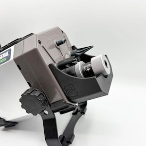 Puede incluir: Un dispositivo Arsenal Center plateado y gris con detalles en negro y una estructura de soporte negra. Cuenta con un componente cilíndrico con borde estriado y un accesorio negro en ángulo. El dispositivo está sobre un fondo blanco.