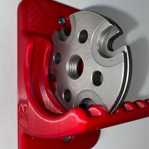 Puede incluir: Un soporte de pared de plástico rojo con un portaherramientas de metal. El portaherramientas tiene un agujero roscado y una serie de ranuras. El soporte de pared tiene el texto "AZ WARE" impreso en él.