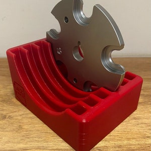 May include: A red plastic holder with five slots for storing metal reloading dies. A single die is shown in the holder.