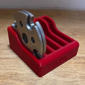 Peut inclure: Un support rouge imprimé en 3D avec le mot "ABL" sur le côté, tenant un disque métallique argenté avec plusieurs trous.