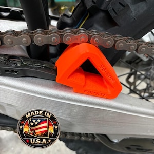 Könnte beinhalten: Ein leuchtend orangefarbener, dreieckiger Motorrad-Kettengleitblock mit dem eingeprägten Text SB-61MM und FOR KTM an der Seite. Das Bauteil ist unter einer metallenen Antriebskette und über einer silbernen Schwinge positioniert und zeigt ein Logo.
