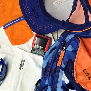 May include: A blue and orange backpack with a camouflage pattern. The backpack has a zippered pocket and a label that says "Property of Jason Reedman". There is also a baseball, a blue baseball cap, a white shirt with an orange pocket, a pair of blue sunglasses, and a red stamp with the word "Trodat" on it.