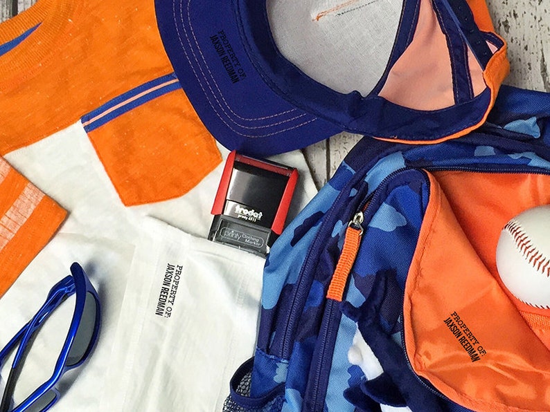 May include: A blue and orange backpack with a camouflage pattern. The backpack has a zippered pocket and a label that says "Property of Jason Reedman". There is also a baseball, a blue baseball cap, a white shirt with an orange pocket, a pair of blue sunglasses, and a red stamp with the word "Trodat" on it.