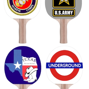 May include: Four different designs of table tennis paddles. The first paddle has a white background with a gold and blue United States Marine Corps logo. The second paddle has a gray background with a black star and the text "U.S. ARMY". The third paddle has a blue background with a white outline of the state of Texas and the text "TEXAS The Lone Star State". The fourth paddle has a white background with a red circle and the text "UNDERGROUND" in blue.