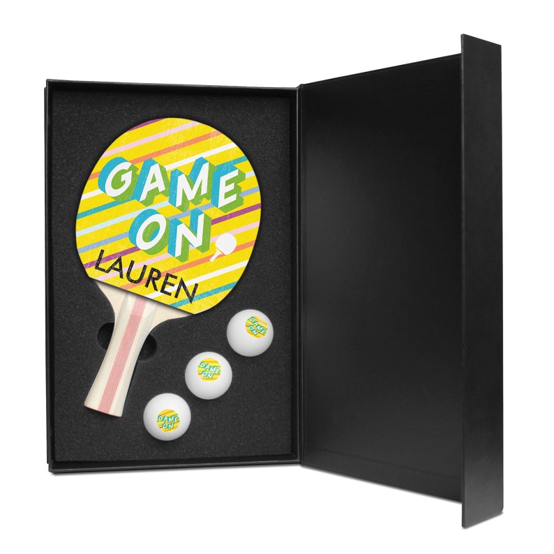 May include: A yellow and blue ping pong paddle with the text "GAME ON" and a personalized name "LAUREN". The paddle is in a black box with three white ping pong balls.