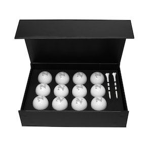 May include: A black box with a hinged lid is open, revealing a set of 12 white golf balls with a black logo and two white golf tees.