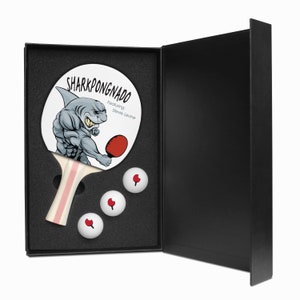 May include: A black box with a white and red ping pong paddle with a cartoon shark design. The paddle has the text "SHARKPONGNADO Featuring: Steve Levine". The box contains three white ping pong balls.