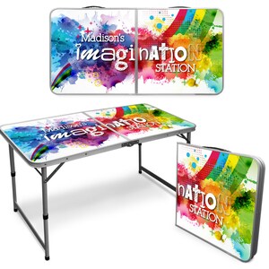 May include: A white folding table with a colorful abstract design and the text "Madison's Imagination Station". The table is made of aluminum and has a black frame. The table is folded up and standing on its side.