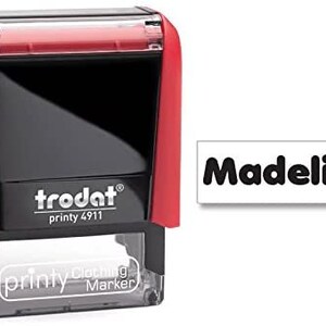 May include: Red and black self-inking stamp with the text "trodat printy 4911" and "printy clothing marker" on the front. The stamp is labeled with the name "Madeline" in black text on a white background.